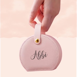 May include: A light pink, round jewelry case with a handle, held by a hand. The case has a gold-coloured clasp and the name "Abbi" in script. The case is approximately 10 cm in diameter.