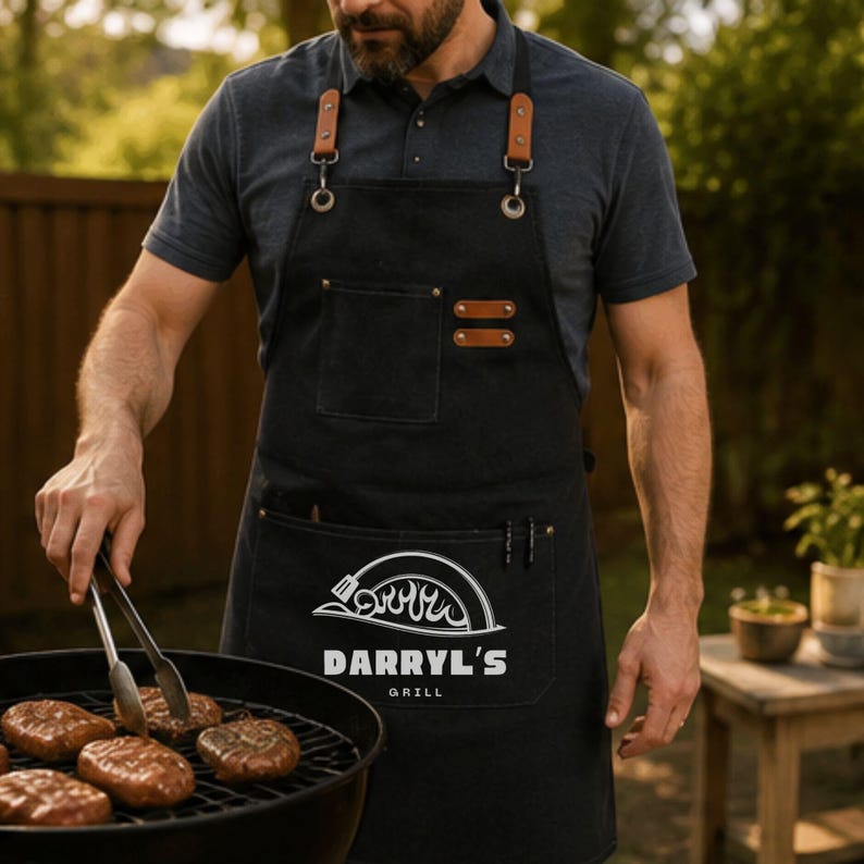 May include: A black apron with brown leather straps and metal hardware, featuring a white graphic of a grill lid with flames and the text DARRYL'S GRILL printed on the front pocket. The apron has a chest pocket and a large lower pocket.
