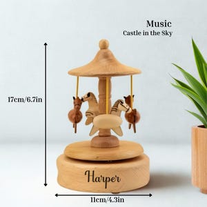 Puede incluir: Una caja de m&uacute;sica de carrusel de madera con cuatro figuras de caballos. La caja de m&uacute;sica mide 17 cm de alto y 11 cm de ancho. El texto "Music Castle in the Sky" est&aacute; encima del carrusel. El nombre "Harper" est&aacute; grabado en la base.