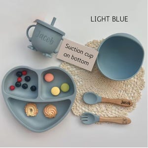 May include: Light blue baby feeding set with a heart-shaped divided plate, bowl, cup with straw, spoon, and fork. The spoon and fork have wooden handles. The cup and spoon are personalized with the name "Jacob".