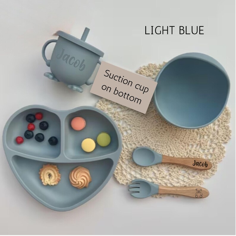 May include: Light blue baby feeding set with a heart-shaped divided plate, bowl, cup with straw, spoon, and fork. The spoon and fork have wooden handles. The cup and spoon are personalized with the name "Jacob".