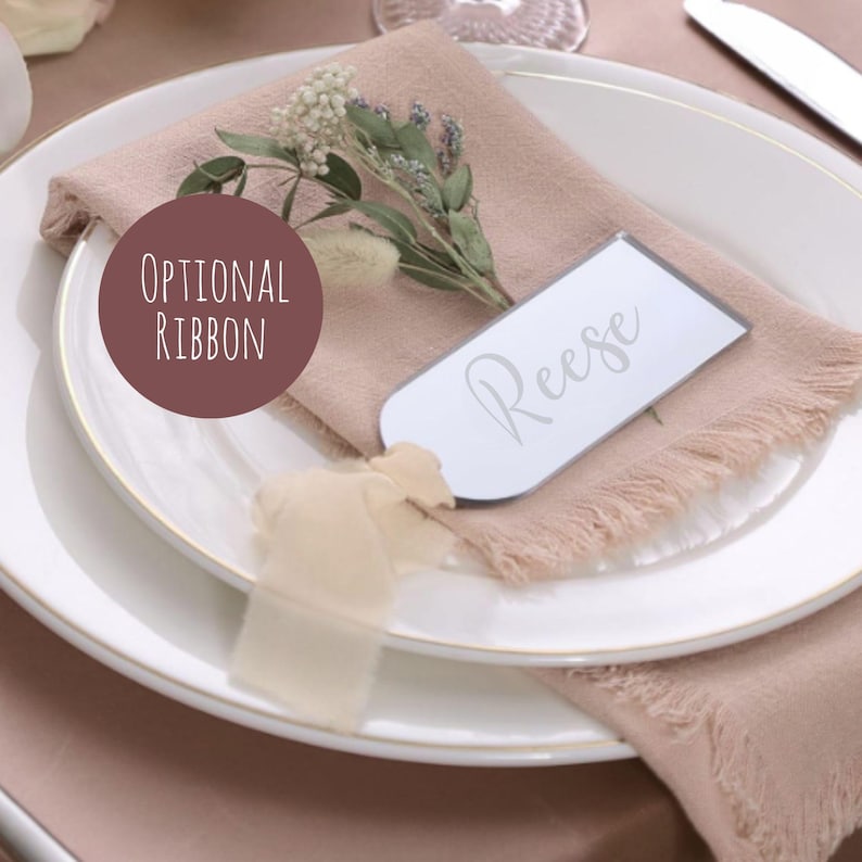 May include: A white plate with a gold rim holds a folded mauve linen napkin. A small sprig of greenery and a white name card that says "Reese" are on the napkin. A ribbon is attached to the name card. A circular graphic says "Optional Ribbon".