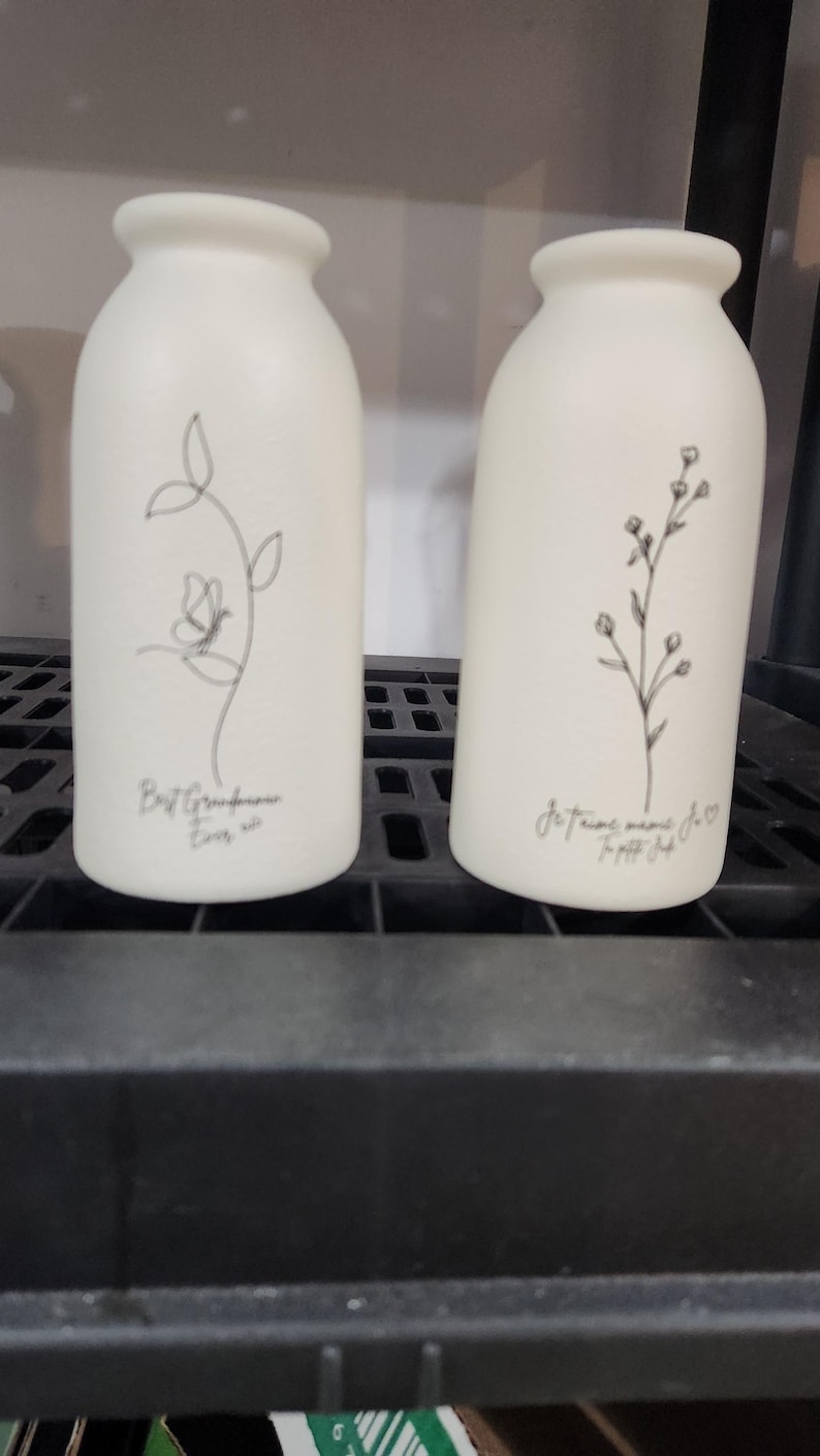 Pu&ograve; includere: Due vasi in ceramica bianca opaca con colli stretti. Il vaso a sinistra presenta un disegno a linee nere di una farfalla su uno stelo con il testo Best Grandmother Ever. Il vaso a destra mostra un disegno a linee nere di un rametto floreale.