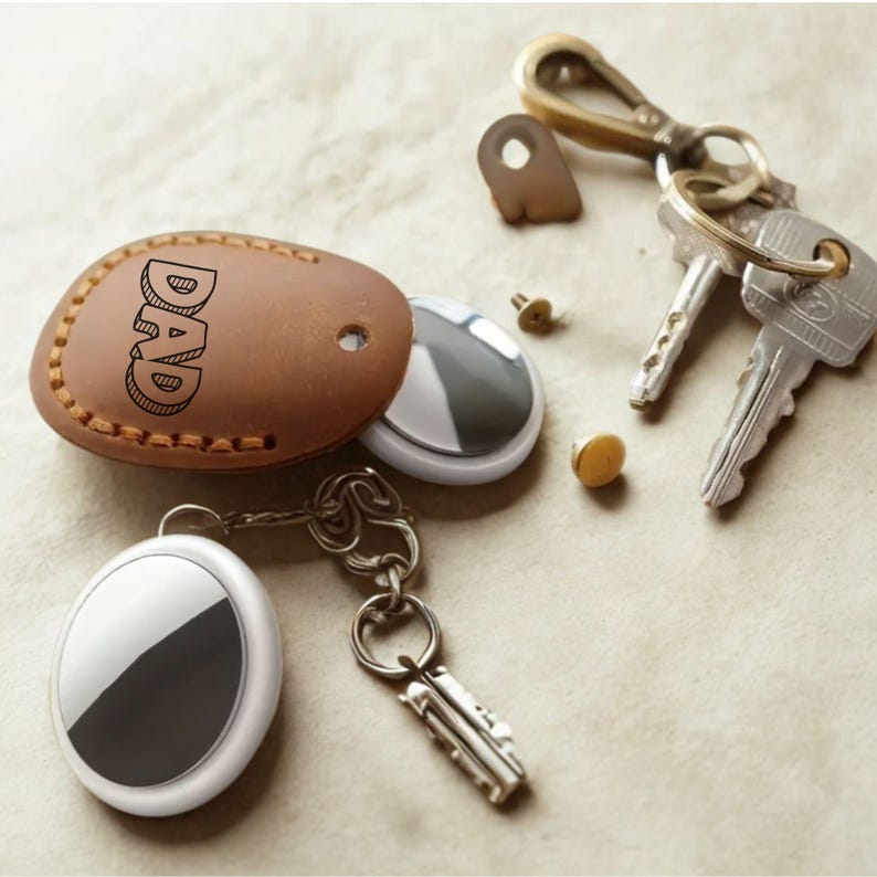 Pu&ograve; includere: Una collezione di chiavi e portachiavi. Un portachiavi in pelle marrone con la scritta "DAD" &egrave; accanto a un dispositivo di localizzazione argento e bianco. Sono visibili anche un fermaglio in ottone e diverse chiavi.