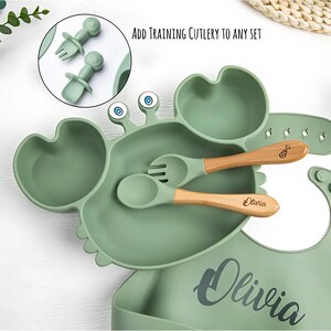 K&ouml;nnte beinhalten: Ein gr&uuml;nes Baby-Set aus Silikon in Krabbenform, bestehend aus einem Teller und einer L&auml;tzchen. Der Teller hat einen Bereich f&uuml;r Lebensmittel und zwei augenf&ouml;rmige Bereiche. Enth&auml;lt eine Gabel und einen L&ouml;ffel mit Holzgriffen. Der Text "Olivia" ist auf dem L&auml;tzchen und dem L&ouml;ffel aufgedruckt.