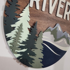 May include: A round wooden wall art piece with the word "RIVER" in white letters. The design features layered trees in shades of green and blue, mountains, and a river. The background is a warm brown wood grain.