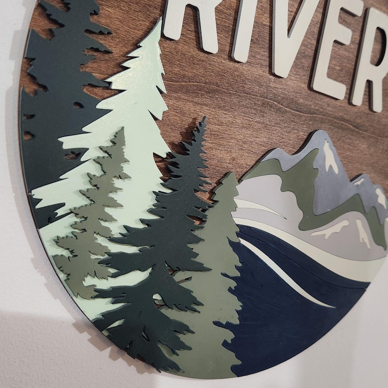 May include: A round wooden wall art piece with the word "RIVER" in white letters. The design features layered trees in shades of green and blue, mountains, and a river. The background is a warm brown wood grain.