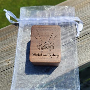 May include: A rectangular wooden box with a dark brown finish, featuring a laser-engraved line art illustration of two hands holding pinky fingers with a small heart above them, and the text Michel and Sydney printed below the illustration.