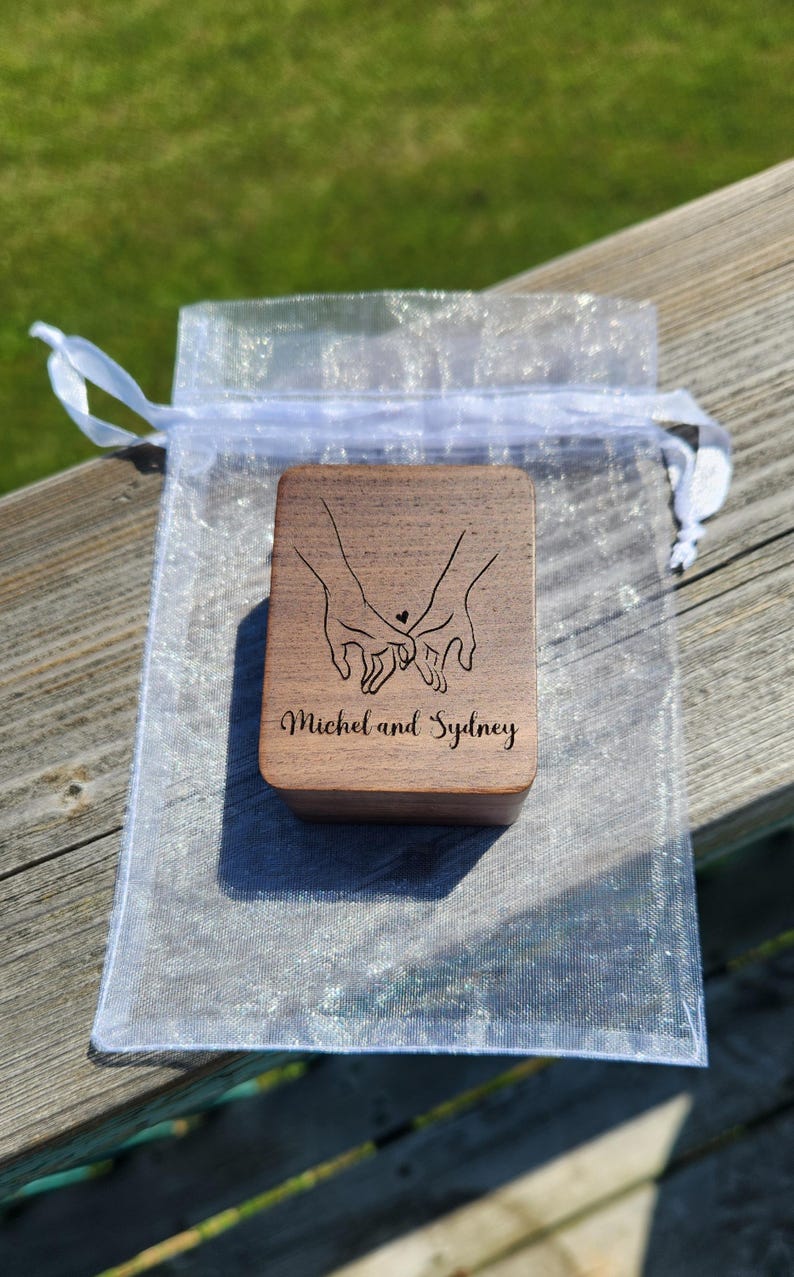 May include: A rectangular wooden box with a dark brown finish, featuring a laser-engraved line art illustration of two hands holding pinky fingers with a small heart above them, and the text Michel and Sydney printed below the illustration.