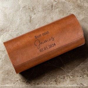 May include: A rectangular tan leather watch roll with a flap closure. The exterior is engraved with the text "Best man", the name "James" in a cursive script, and the date "02.03.2024" in a clean, dark, sans-serif font.