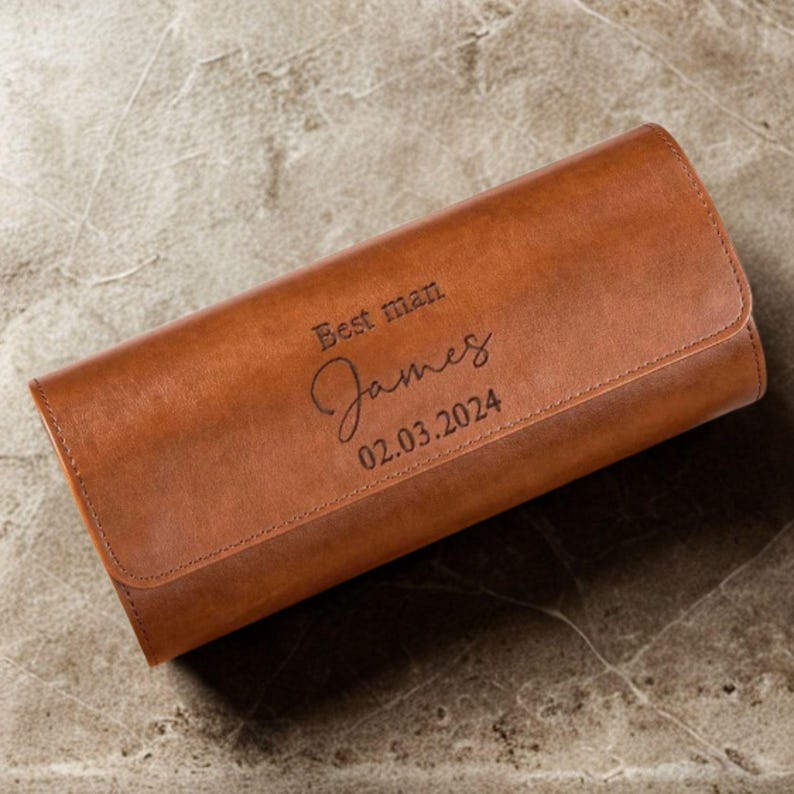 May include: A rectangular tan leather watch roll with a flap closure. The exterior is engraved with the text "Best man", the name "James" in a cursive script, and the date "02.03.2024" in a clean, dark, sans-serif font.