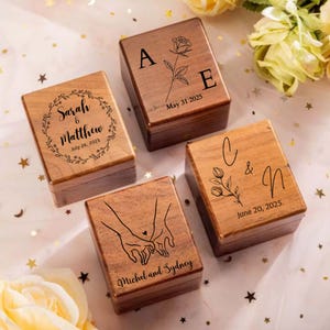May include: Four square wooden boxes with hinged lids, each featuring unique laser-engraved designs. The boxes display names, dates, and illustrations including a rose, a floral wreath, a botanical sprig, and two hands holding each other.