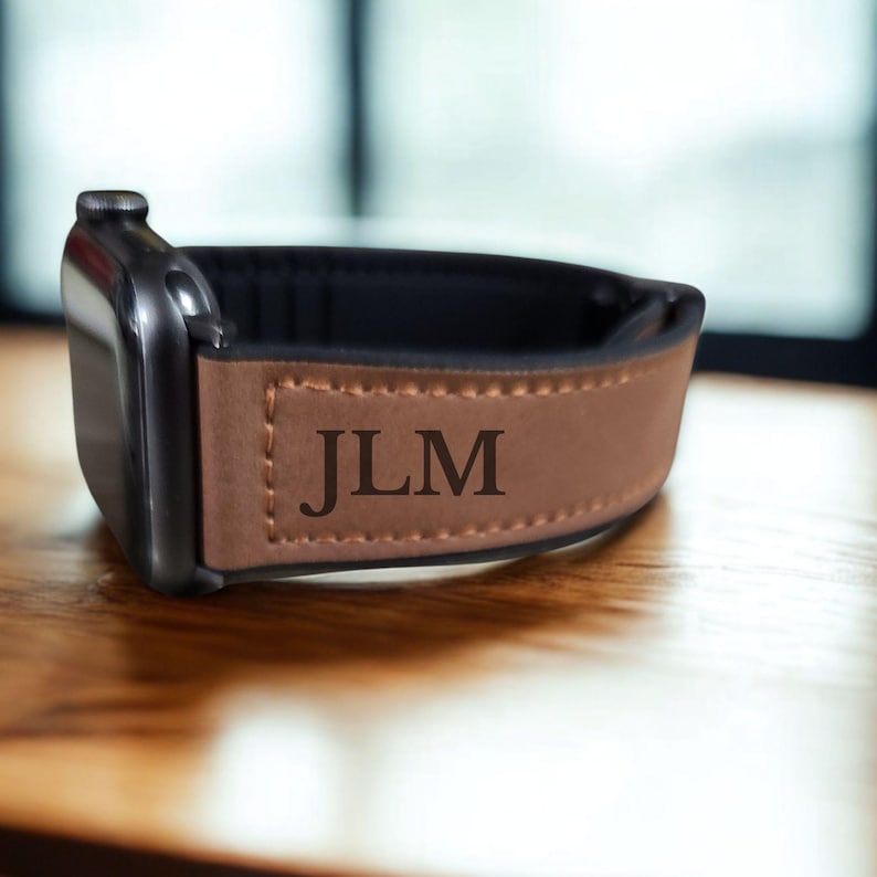 Pu&ograve; includere: Un cinturino per orologio in pelle marrone con le iniziali "JLM" incise sul davanti. Il cinturino &egrave; attaccato a uno smartwatch nero. Il cinturino &egrave; realizzato in pelle marrone chiaro e ha un interno nero.