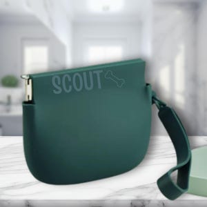 K&ouml;nnte beinhalten: Eine teal-farbene Tasche mit dem Wort "SCOUT" und einer Knochengrafik auf der Vorderseite. Die Tasche hat einen kleinen Griff und einen Metallverschluss. Der Hintergrund ist unscharf.