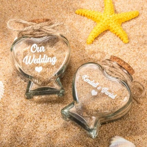 May include: Two heart-shaped glass jars filled with sand, each topped with a cork and twine. One jar features the white text Our Wedding with a heart icon, while the other displays the white text Kendall e Daniel with a heart icon.