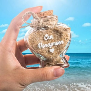 May include: A heart-shaped glass bottle filled with light brown sand, featuring a cork stopper tied with a jute twine bow. The front of the glass is printed with the white text Our Honeymoon and a small white heart icon below the lettering.