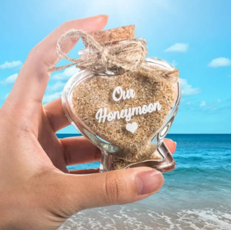 May include: A heart-shaped glass bottle filled with light brown sand, featuring a cork stopper tied with a jute twine bow. The front of the glass is printed with the white text Our Honeymoon and a small white heart icon below the lettering.