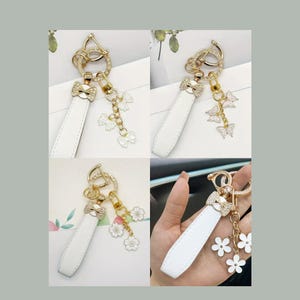 Op de afbeelding: Een verzameling witte leren sleutelhangers met goudkleurige accenten. Elke sleutelhanger heeft een strikvormige bedel met strass-steentjes en bungelende bedels, waaronder strikken, vlinders en bloemen. Weergegeven in een collage.