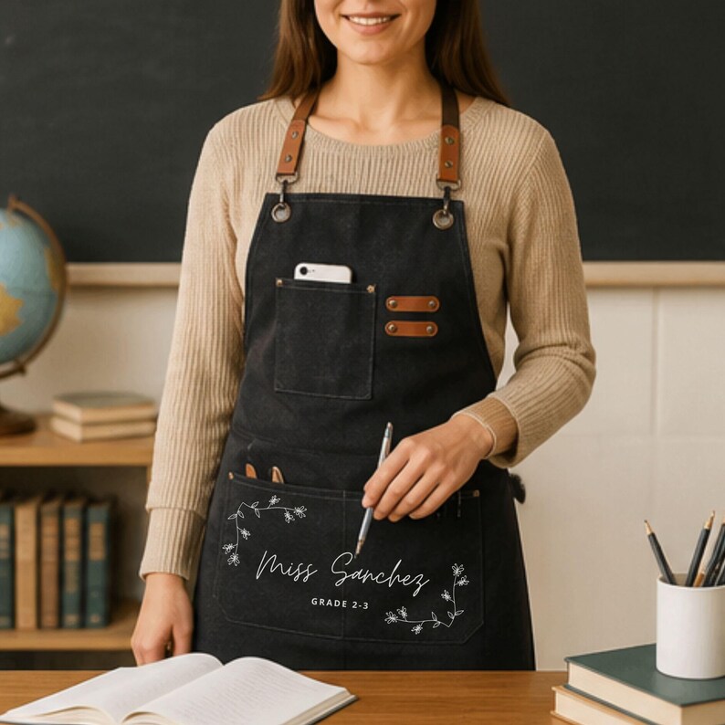 May include: A black canvas apron with brown leather straps, metal hardware, and multiple pockets. The front pocket features white floral line art, the text "Miss Sanchez", and "GRADE 2-3" printed in a clean, white, sans-serif font.