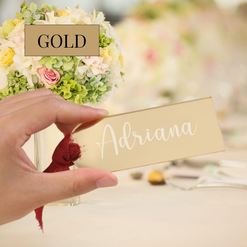 Bridal shower table setting featuring acrylic name signs atop napkins on classic china plates, with white roses and glassware in the background, used as guest name tags and keepsake gifts for attendees.