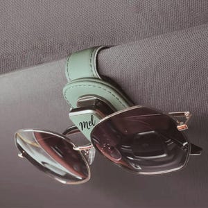 K&ouml;nnte beinhalten: Ein salbeigr&uuml;ner Sonnenbrillenhalter aus Leder f&uuml;r die Sonnenblende im Auto mit einem Metallclip-Mechanismus und dem in schwarzer Schreibschrift aufgedruckten Namen mel. Der Halter ist an einer grauen Stoffblende befestigt und h&auml;lt eine Sonnenbrille.