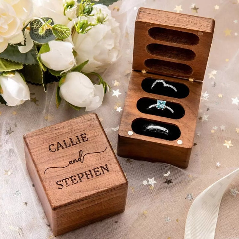 May include: A dark wood ring box with a hinged lid, engraved with the names CALLIE and STEPHEN. The open box features three horizontal slots containing three silver-toned rings, including one with a square blue stone and two plain bands.