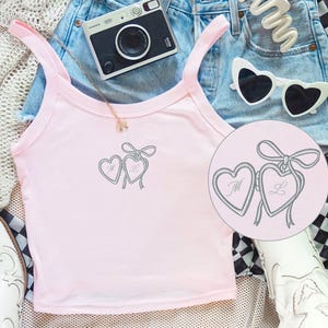 May include: A light pink tank top with a heart design, paired with denim shorts, heart-shaped sunglasses, and a black and white checkered scarf. A small camera and white boots complete the casual outfit.
