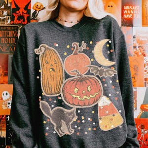 May include: A dark heather gray crewneck sweatshirt featuring a graphic print of a tall pumpkin, a round jack-o'-lantern, a crescent moon, a bat, a black cat, and a piece of candy corn, all surrounded by small colorful dots in a festive pattern.