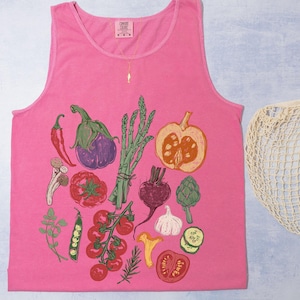 May include: A vibrant pink vest top featuring a vegetable illustration with a red chilli, aubergine, asparagus, and tomatoes. A beige string bag is next to the vest top.