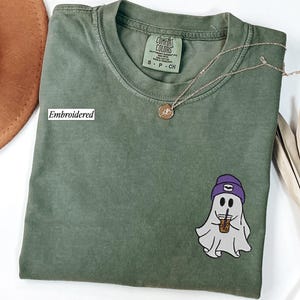 May include: A green Comfort Colors t-shirt with an embroidered ghost graphic. The ghost wears a purple beanie and holds a coffee cup. The shirt has a Comfort Colors tag and a gold necklace. The word 'Embroidered' is printed on the shirt.