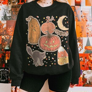May include: A black crewneck sweatshirt featuring a graphic print of a tall orange jack-o'-lantern, a round jack-o'-lantern, a crescent moon, a bat, a black cat, and a piece of candy corn, all surrounded by small multicolored confetti dots.