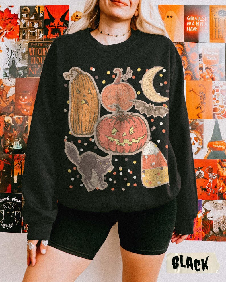 May include: A black crewneck sweatshirt featuring a graphic print of a tall orange jack-o'-lantern, a round jack-o'-lantern, a crescent moon, a bat, a black cat, and a piece of candy corn, all surrounded by small multicolored confetti dots.