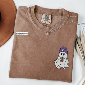 May include: A brown t-shirt with an embroidered ghost design wearing a purple beanie and holding a coffee cup. The shirt has a crew neck and a small tag. A silver necklace with a pendant is also visible.