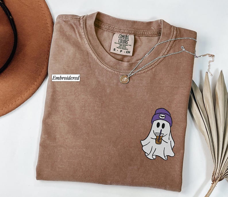 May include: A brown t-shirt with an embroidered ghost design wearing a purple beanie and holding a coffee cup. The shirt has a crew neck and a small tag. A silver necklace with a pendant is also visible.