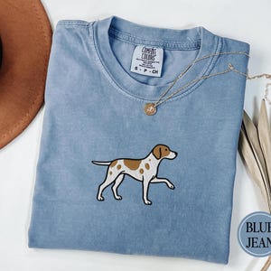 Puede incluir: Camiseta azul claro con un diseño de perro bordado. El perro es blanco con manchas marrones. Un collar dorado con un colgante descansa sobre la camiseta. Un sombrero marrón y plantas secas están en el fondo. La camiseta tiene una etiqueta que dice "Comfort Colors".