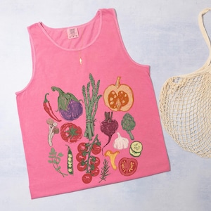May include: A vibrant pink vest top featuring a detailed print of assorted vegetables such as tomatoes, aubergine, and asparagus. A cream-colored mesh shopping bag is positioned beside the top. The top is made of soft material.