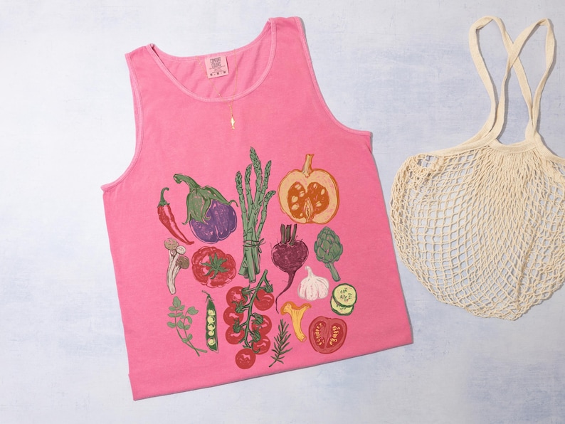 May include: A vibrant pink vest top featuring a detailed print of assorted vegetables such as tomatoes, aubergine, and asparagus. A cream-colored mesh shopping bag is positioned beside the top. The top is made of soft material.