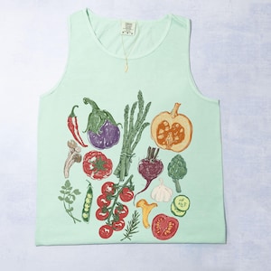 May include: A pale green vest top with a detailed illustration of vegetables. The design includes a red chilli, aubergine, tomato, pumpkin, and asparagus. The artwork is in a hand-drawn style.