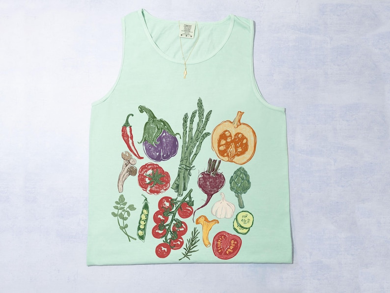 May include: A pale green vest top with a detailed illustration of vegetables. The design includes a red chilli, aubergine, tomato, pumpkin, and asparagus. The artwork is in a hand-drawn style.