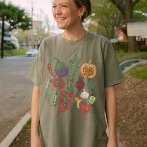 May include: Olive green t-shirt featuring a colorful illustration of various vegetables, including tomatoes, eggplant, and a pumpkin. The design is printed on a short-sleeved, crew neck tee. The shirt is worn by a person.