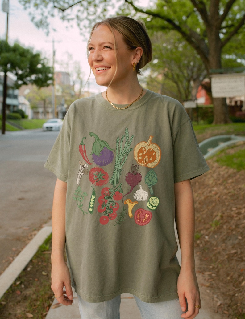 May include: Olive green t-shirt featuring a colorful illustration of various vegetables, including tomatoes, eggplant, and a pumpkin. The design is printed on a short-sleeved, crew neck tee. The shirt is worn by a person.