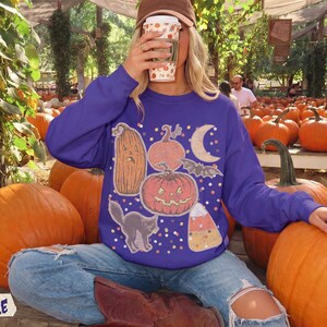 May include: A purple long-sleeve sweatshirt featuring a graphic print of a tall pumpkin, a round jack-o'-lantern, a crescent moon, a bat, a black cat, and a piece of candy corn, all surrounded by small colorful dots on the front of the garment.