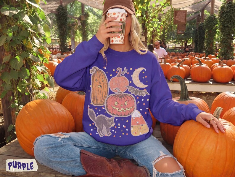 May include: A purple long-sleeve sweatshirt featuring a graphic print of a tall pumpkin, a round jack-o'-lantern, a crescent moon, a bat, a black cat, and a piece of candy corn, all surrounded by small colorful dots on the front of the garment.