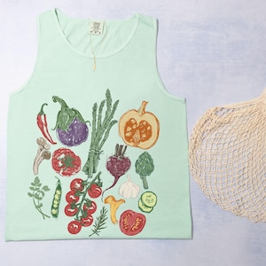 May include: A pale green vest top featuring a vibrant illustration of vegetables such as tomatoes, aubergine, and asparagus. A beige mesh tote bag is next to the top. A gold necklace hangs from the neckline.