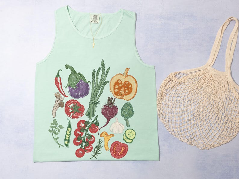 May include: A pale green vest top featuring a vibrant illustration of vegetables such as tomatoes, aubergine, and asparagus. A beige mesh tote bag is next to the top. A gold necklace hangs from the neckline.