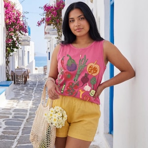 May include: A woman in a pink vest top embroidered with vegetables, and yellow shorts. She holds a beige mesh bag with white flowers. The background shows a white street with blue details and pink flowers, reminiscent of a Mediterranean village.