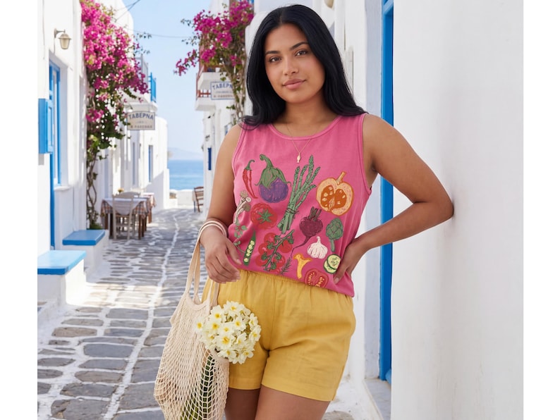 May include: A woman in a pink vest top embroidered with vegetables, and yellow shorts. She holds a beige mesh bag with white flowers. The background shows a white street with blue details and pink flowers, reminiscent of a Mediterranean village.