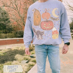 May include: A light grey crewneck sweatshirt featuring a graphic print of Halloween-themed icons including a tall gourd, a round jack-o'-lantern, a crescent moon, a flying bat, a black cat, and a piece of candy corn, all surrounded by colorful dots.