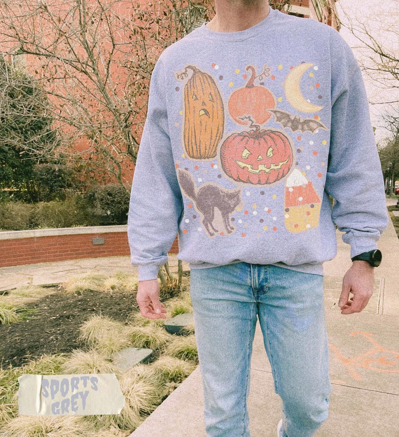 May include: A light grey crewneck sweatshirt featuring a graphic print of Halloween-themed icons including a tall gourd, a round jack-o'-lantern, a crescent moon, a flying bat, a black cat, and a piece of candy corn, all surrounded by colorful dots.