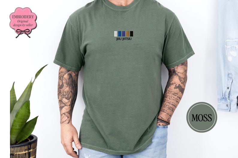 May include: Olive green t-shirt with the word "JIU JITSU" embroidered in black below a row of coloured rectangles. The shirt is short-sleeved and has a relaxed fit. The model has tattoos on their arms.